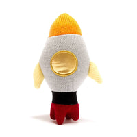 A knitted plush toy shaped like a rocket ship, shown from the back on a clean white background. The rocket features a white body, a yellow-orange pointed nose, a yellow circular window, light yellow wings, a black band, and red feet. Perfect to combine with one of ur premature baby clothes. We also provide gift shipping