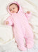 A baby lying on a white textured blanket wearing a pink hooded footed sleeper with small dark polka dots.