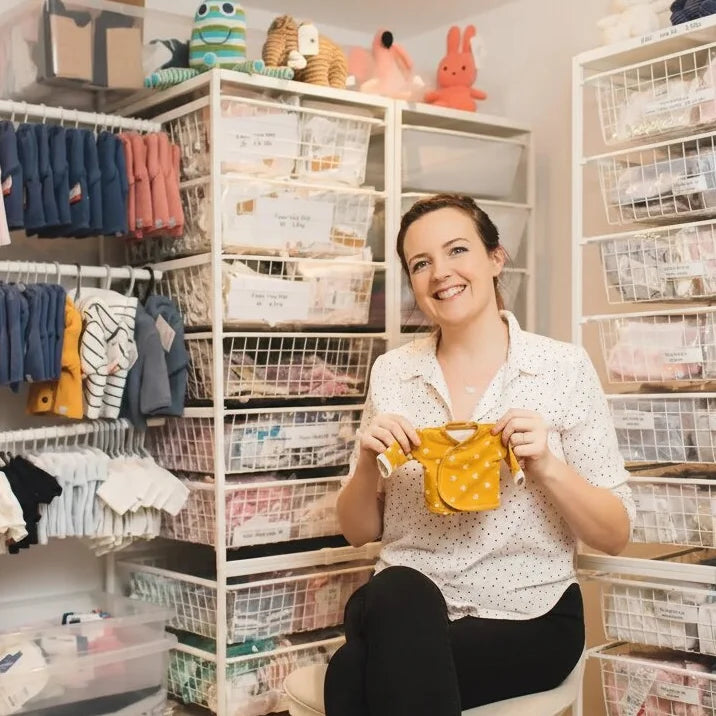Woman holding prem baby clothing in a storage room with shelves and bins.
