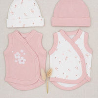 A baby clothing set laid flat on a light beige fabric background, including a white knit beanie with tiny pink floral prints, a solid light pink beanie, a pink wrap vest with white floral embroidery, and a white wrap vest with pink trim and small pink floral prints. A small dried decorative stem sits between the garments.