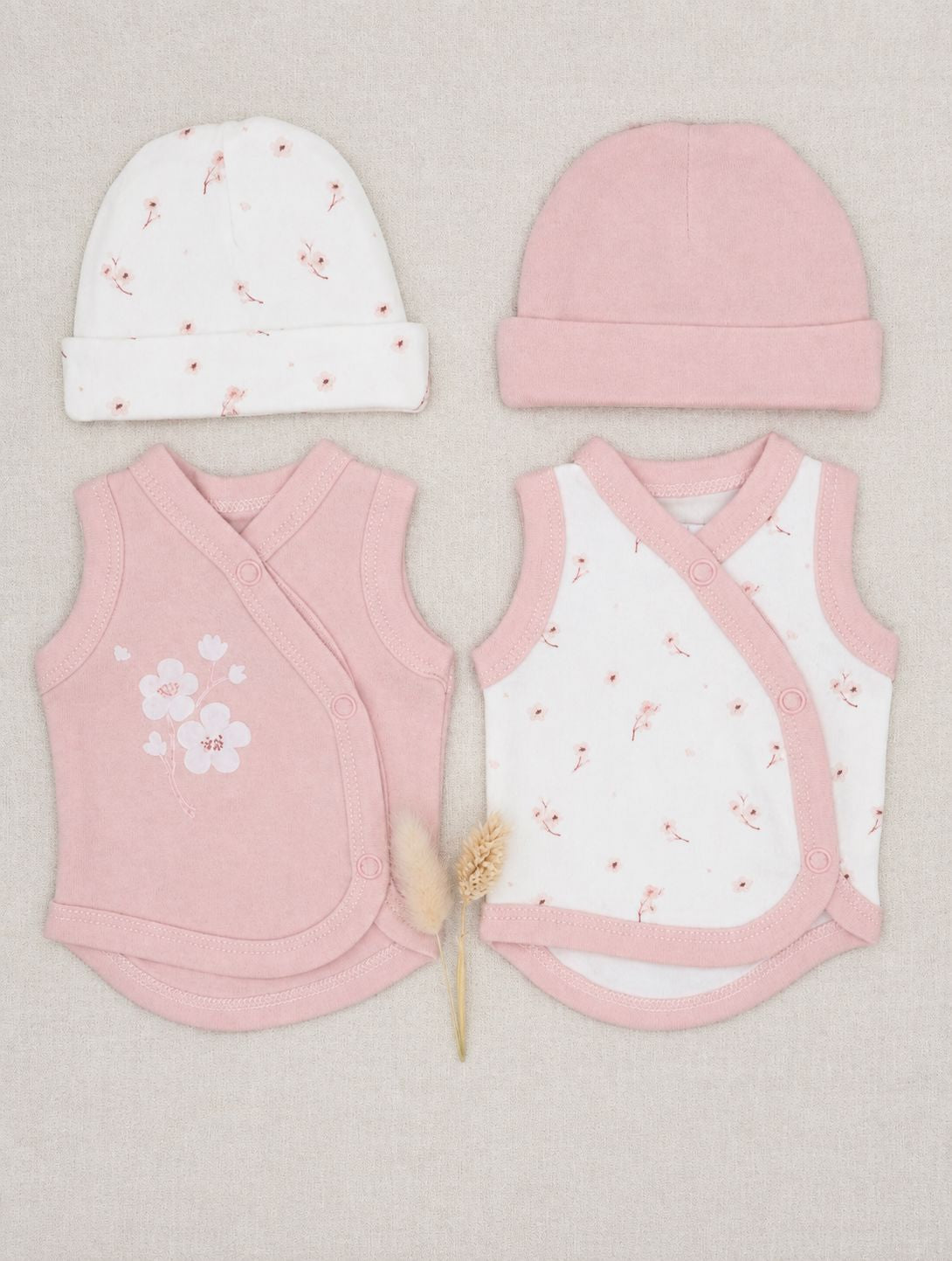 A baby clothing set laid flat on a light beige fabric background, including a white knit beanie with tiny pink floral prints, a solid light pink beanie, a pink wrap vest with white floral embroidery, and a white wrap vest with pink trim and small pink floral prints. A small dried decorative stem sits between the garments.