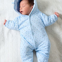 A sleeping newborn baby wearing a light blue hooded one-piece snowsuit with small dark polka dots, lying on a textured white blanket.