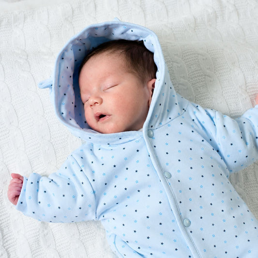 A sleeping newborn baby wearing a light blue hooded onesie with small blue and black polka dots, lying on a white textured blanket.