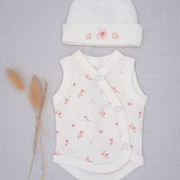 A baby outfit set featuring a white sleeveless wrap bodysuit and matching beanie hat, both decorated with a delicate pink floral pattern, laid flat on a light gray textured background next to dried pampas grass.
