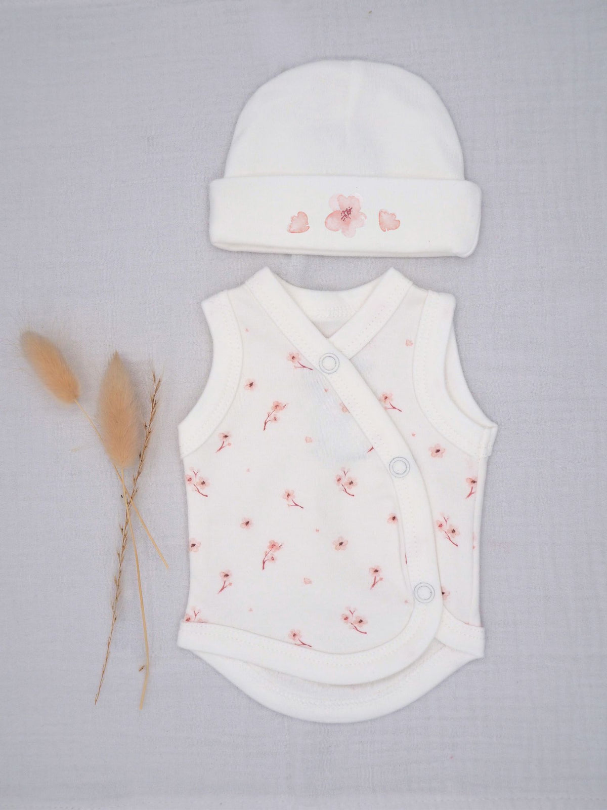 A baby outfit set featuring a white sleeveless wrap bodysuit and matching beanie hat, both decorated with a delicate pink floral pattern, laid flat on a light gray textured background next to dried pampas grass.