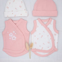 A baby clothing set laid flat on a white textured fabric background, including a pink knit hat, a white knit hat with small pink floral prints, a pink wrap vest, and a white wrap vest with pink trim and tiny pink floral prints. A small dried flower stem lies between the garments.