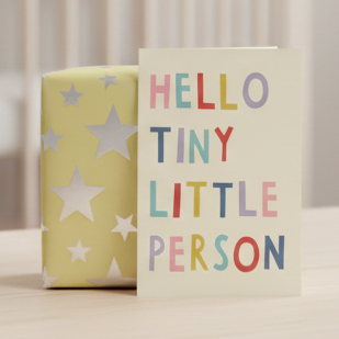 A cream greeting card with the phrase 'HELLO TINY LITTLE PERSON' in multicolored letters, standing next to a pale yellow gift box with white star patterns on a light wooden surface.