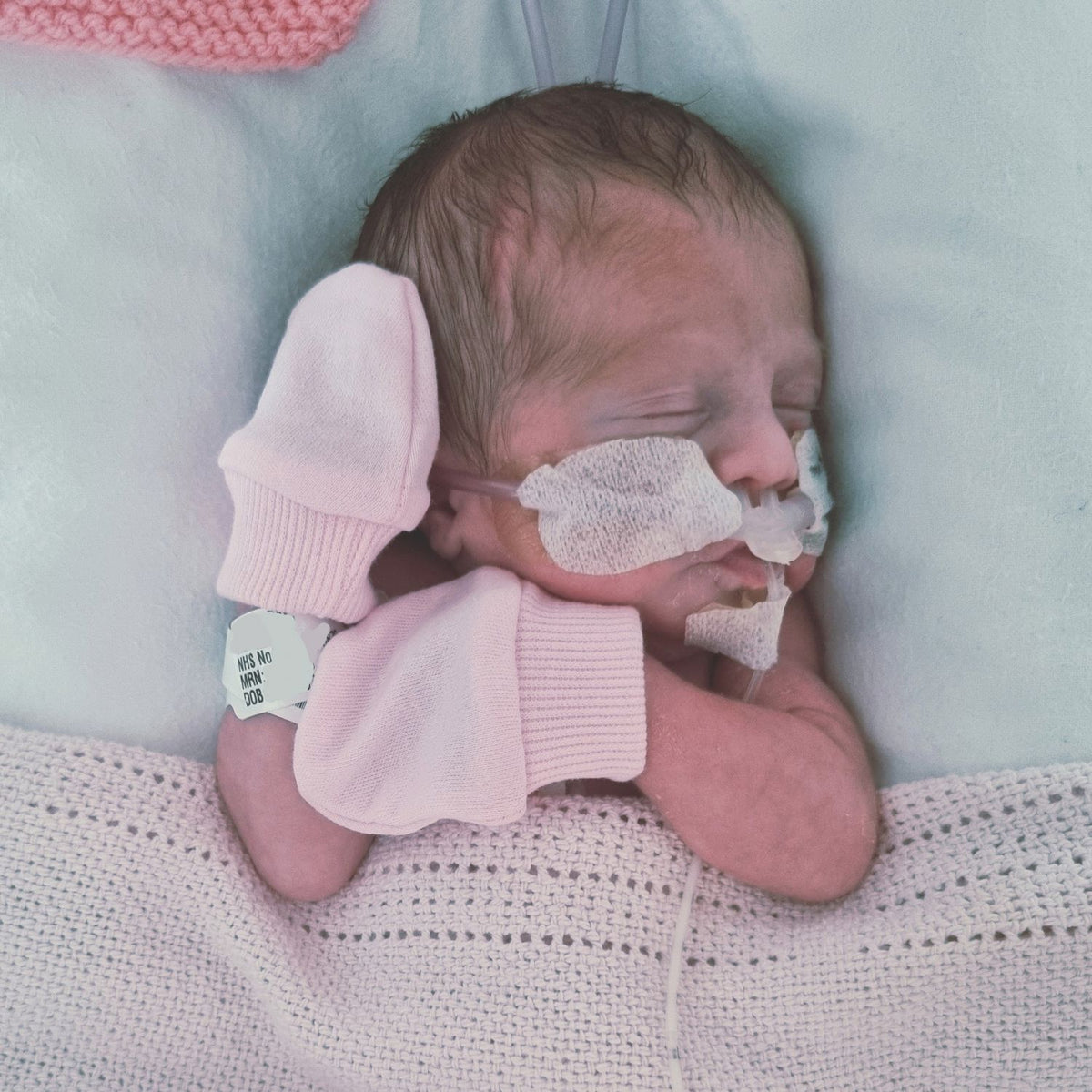 A newborn baby sleeping peacefully, swaddled in a light pink knit blanket and wearing a matching pair of pale pink knit booties, with a small white tag visible on one bootie.