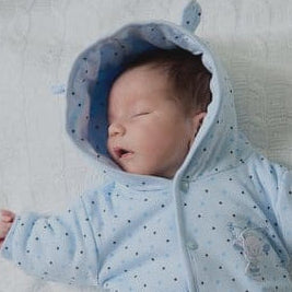 Newborn baby wearing a blue onesie with a hood on a white blanket.